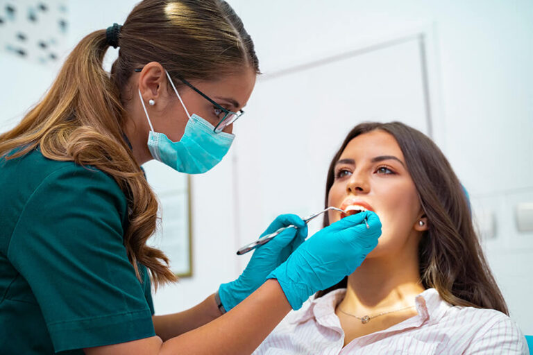 The image shows a dental professional in a green uniform and blue gloves examining a patient's mouth during a dental checkup. The patient, seated in a dental chair, is wearing a white shirt with a necklace. The setting clearly indicates a dental clinic.