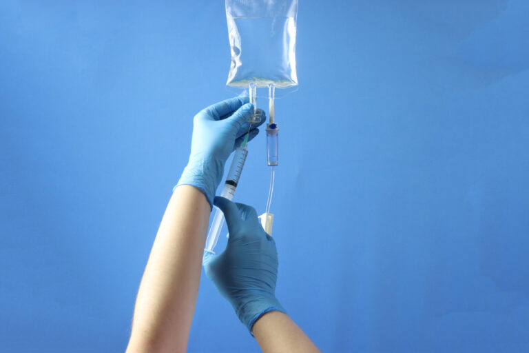 Gloved hands adjusting a syringe connected to an intravenous (IV) line, with a transparent fluid bag containing medical solution in the background. This image depicts a common medical procedure in healthcare settings.