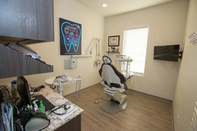 A dental office featuring a centrally positioned chair equipped with a light and essential instruments. Nearby, a desk holds a computer and dental supplies, while a wall poster illustrates a tooth with colorful graphics and text.