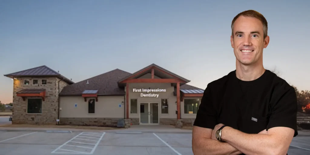 A building labeled 'First Impressions Dentistry' with a modern stone and stucco exterior, multiple windows, and a pitched roof. A person in a black shirt stands with arms crossed near the parking lot in front.